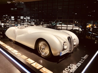 A vintage white sports car displayed in a dimly lit showroom with reflective black walls. The car has a sleek, aerodynamic body with a rounded front, prominent headlights, and a clearly visible license plate. Information and images about the car are placed alongside it on a panel.