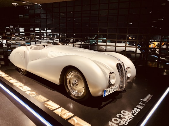 A vintage white sports car displayed in a dimly lit showroom with reflective black walls. The car has a sleek, aerodynamic body with a rounded front, prominent headlights, and a clearly visible license plate. Information and images about the car are placed alongside it on a panel.