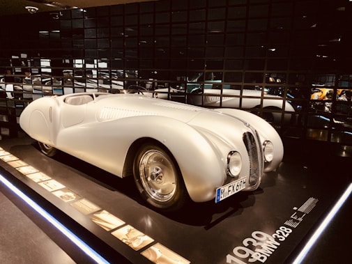 A vintage white sports car displayed in a dimly lit showroom with reflective black walls. The car has a sleek, aerodynamic body with a rounded front, prominent headlights, and a clearly visible license plate. Information and images about the car are placed alongside it on a panel.