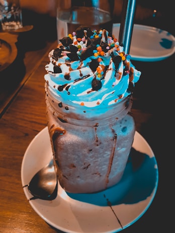 A creamy dessert drink served in a mason jar, topped with whipped cream, chocolate sauce, and colorful sprinkles. The drink is placed on a round white plate with a metal straw and a spoon beside it.