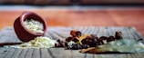 A small earthen pot tipped over on a wooden surface spills uncooked rice. Surrounding the rice are various spices including cinnamon sticks, cloves, star anise, and bay leaves, creating a rustic and aromatic tableau.