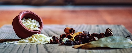 A small earthen pot tipped over on a wooden surface spills uncooked rice. Surrounding the rice are various spices including cinnamon sticks, cloves, star anise, and bay leaves, creating a rustic and aromatic tableau.