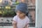 A young child with a thoughtful expression is wearing a patterned blue turban and a light pink shirt with delicate lace detailing. The background includes a blurred outdoor setting with greenery and some architectural elements.