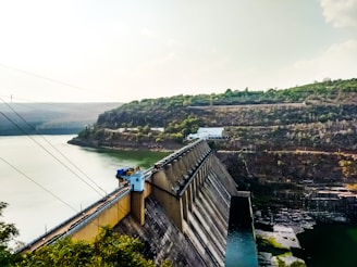 A large dam stretches across a river, holding back a significant volume of water. The structure is massive and constructed from concrete, with various industrial components such as towers and cables. Steep hills covered with sparse vegetation flank the riverbanks, while the sky above is a soft gradient of blue and white, suggesting a partly cloudy day.