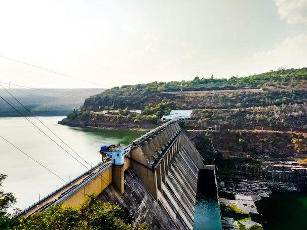 A large dam stretches across a river, holding back a significant volume of water. The structure is massive and constructed from concrete, with various industrial components such as towers and cables. Steep hills covered with sparse vegetation flank the riverbanks, while the sky above is a soft gradient of blue and white, suggesting a partly cloudy day.