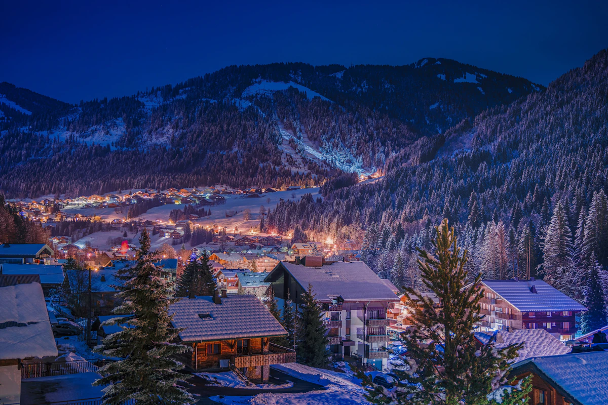 Small mountain town glowing at dusk below snow-covered slopes and pine forest