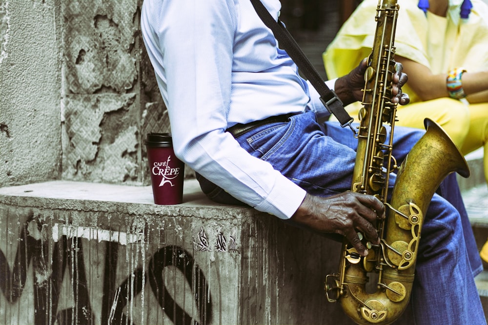 man in white long sleeve shirt and blue denim jeans sitting on concrete wall playing saxophone