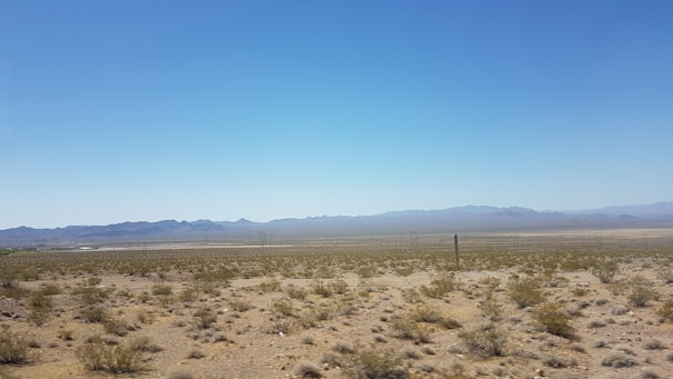A vast desert landscape stretches into the distance with sparse vegetation scattered across the sandy ground. Low mountain ranges are visible on the horizon under a clear, blue sky. The scene is mostly flat, with some distant utility poles indicating human presence.