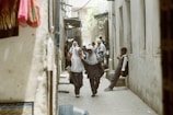 Two schoolchildren in uniforms and hijabs walk down a narrow street, smiling and engaged in conversation, while another child stands against a wall holding a drink. The passage is flanked by walls and the scene appears to be in a bustling area with more people visible in the background.