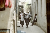 Two schoolchildren in uniforms and hijabs walk down a narrow street, smiling and engaged in conversation, while another child stands against a wall holding a drink. The passage is flanked by walls and the scene appears to be in a bustling area with more people visible in the background.