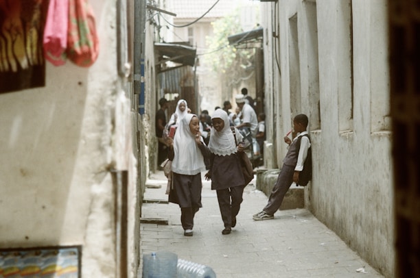 Two schoolchildren in uniforms and hijabs walk down a narrow street, smiling and engaged in conversation, while another child stands against a wall holding a drink. The passage is flanked by walls and the scene appears to be in a bustling area with more people visible in the background.