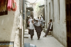 Two schoolchildren in uniforms and hijabs walk down a narrow street, smiling and engaged in conversation, while another child stands against a wall holding a drink. The passage is flanked by walls and the scene appears to be in a bustling area with more people visible in the background.