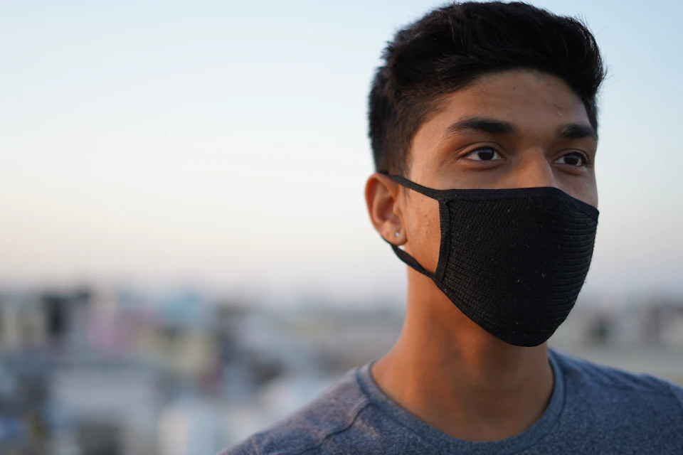 In the image, a young man is standing against a backdrop of a city skyline. He's wearing a black face mask that covers his entire face and extends down to his neck. The mask has a mesh pattern on it, allowing for some airflow while still providing protection from airborne particles or germs. The man appears to be looking off into the distance, possibly lost in thought or observing something happening beyond him.