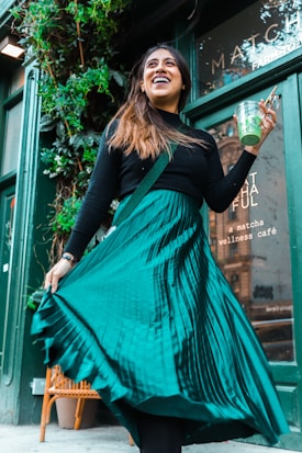 A woman wearing a green pleated skirt and a black top is smiling while holding a cup with a green beverage. She is standing in front of a café with green walls and foliage growing around the entrance.