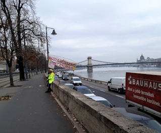 A person wearing a bright yellow jacket is standing on the sidewalk next to a busy street, holding a banner in Hungarian. The street runs parallel to a river, across which a large suspension bridge can be seen. The sky is overcast, and trees without leaves line the sidewalk.