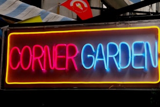 Bright neon sign displaying the words 'Corner Garden' in vibrant red and blue colors. The letters are outlined with a yellow hue, providing a striking contrast. The sign is set against a dark background, highlighting the neon glow.