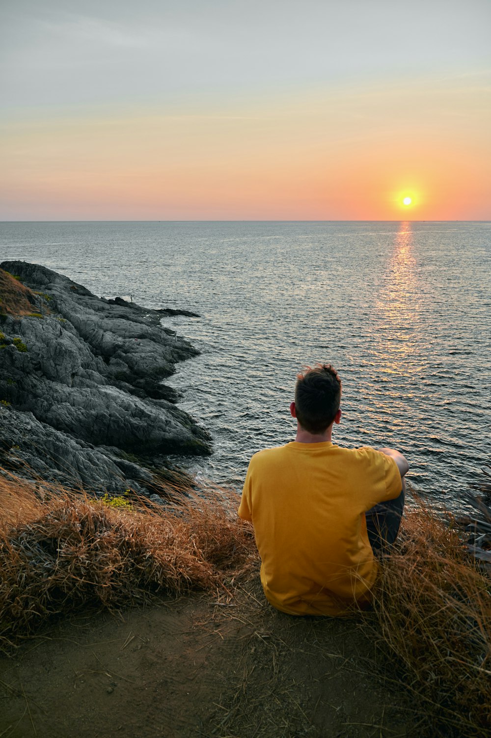 man in yellow shirt sitting on rock near body of water during sunset