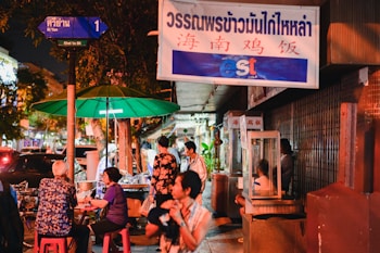 A bustling street food scene at night featuring a group of people sitting and eating around a small table. The area is illuminated by streetlights and a green umbrella. A sign in both Thai and Chinese is prominently displayed above, indicating the presence of a food establishment. The atmosphere is lively with pedestrians and diners interacting.