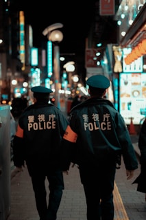 Two police officers are walking down a busy city street at night. They are wearing uniforms with 'POLICE' clearly visible on their backs. The street is illuminated by bright neon signs and streetlights, with a bustling atmosphere created by blurred lights and indistinct figures in the background.