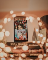 A stack of books with a smartphone resting against them, displaying an image of a person holding a book titled 'How to Be a Bawse' by Lilly Singh. The background of the smartphone image is adorned with colorful flowers. Warm, blurred lights create a soft bokeh effect around the scene, adding a cozy and inviting atmosphere.