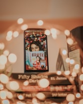 A stack of books with a smartphone resting against them, displaying an image of a person holding a book titled 'How to Be a Bawse' by Lilly Singh. The background of the smartphone image is adorned with colorful flowers. Warm, blurred lights create a soft bokeh effect around the scene, adding a cozy and inviting atmosphere.