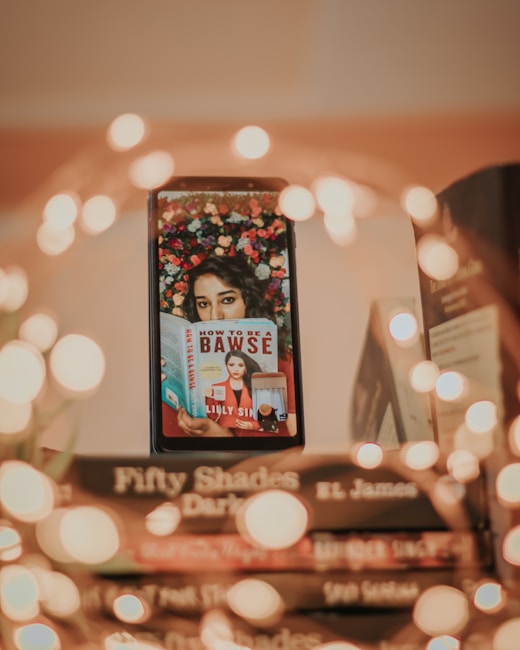 A stack of books with a smartphone resting against them, displaying an image of a person holding a book titled 'How to Be a Bawse' by Lilly Singh. The background of the smartphone image is adorned with colorful flowers. Warm, blurred lights create a soft bokeh effect around the scene, adding a cozy and inviting atmosphere.