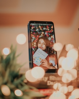 A smartphone displays the image of a woman with curly hair set against a vibrant floral background. She holds a book titled 'the sun and her flowers' with an intricate floral design. Soft, warm lighting creates a bokeh effect, with lights and a blurred leafy plant in the foreground.