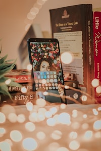 A mobile phone displays a woman's face surrounded by flowers on its screen, placed on a surface with light bokeh effects in the foreground. Nearby, several books are stacked, including titles like 'The Diary of a Young Girl' and 'Little Women'. A small plant is partially visible to the left.