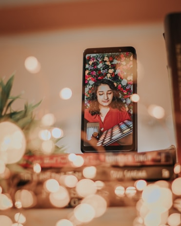 A smartphone is placed upright on a stack of books, displaying a video call or a streaming session with a woman. She is wearing a red top and holding an eyeshadow palette while surrounded by a vibrant floral background. In the foreground, there are blurred lights that create a bokeh effect and some greenery is visible to the side.