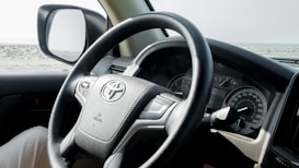 A close-up view of a car's interior, focusing on the steering wheel which has a leather texture and features the Toyota logo. The dashboard displays various dials and gauges, and the side mirror reflects the outdoor landscape, suggesting the car is parked in an open, possibly desert area.