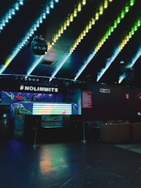 A dimly lit nightclub interior with colorful LED lights on the ceiling creating a vibrant pattern. A DJ booth with graffiti-style art sits under a sign that reads #NOLIMMITS. A colorful light display is on the front of the booth. The surrounding area is dark, with seating arrangements visible in the background.