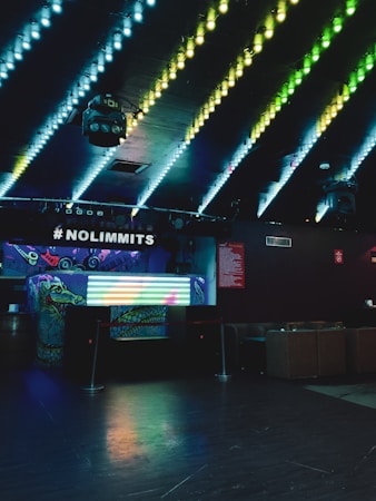A dimly lit nightclub interior with colorful LED lights on the ceiling creating a vibrant pattern. A DJ booth with graffiti-style art sits under a sign that reads #NOLIMMITS. A colorful light display is on the front of the booth. The surrounding area is dark, with seating arrangements visible in the background.