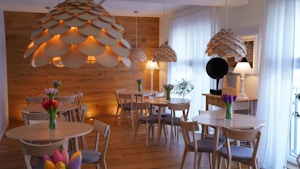 A cozy dining area with wooden walls and floors, featuring several round tables and matching chairs. Large, decorative pendant lights hang from the ceiling, resembling scales or petals illuminated warmly. Each table has a vase of tulips in various colors. Sheer white curtains cover the windows, allowing soft natural light to enter. A side table has a lamp and potted plant.