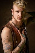 A person with a unique style, featuring curly blond hair, a nose ring, and beard. Tattoos are visible on the arms, including a floral and geometric design. Wearing a sleeveless brown leather vest and multiple necklaces, one of which is blue.