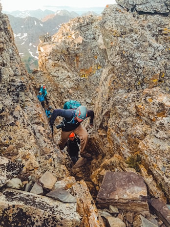 A group of climbers in rugged outdoor gear navigate a narrow, rocky crevice on a mountain. The scene is set amid large, jagged boulders, with some snow-capped peaks visible in the background. Each climber wears helmets and backpacks, emphasizing the challenging and adventurous nature of their ascent.