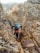 A group of climbers in rugged outdoor gear navigate a narrow, rocky crevice on a mountain. The scene is set amid large, jagged boulders, with some snow-capped peaks visible in the background. Each climber wears helmets and backpacks, emphasizing the challenging and adventurous nature of their ascent.