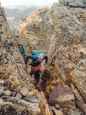 A group of climbers in rugged outdoor gear navigate a narrow, rocky crevice on a mountain. The scene is set amid large, jagged boulders, with some snow-capped peaks visible in the background. Each climber wears helmets and backpacks, emphasizing the challenging and adventurous nature of their ascent.
