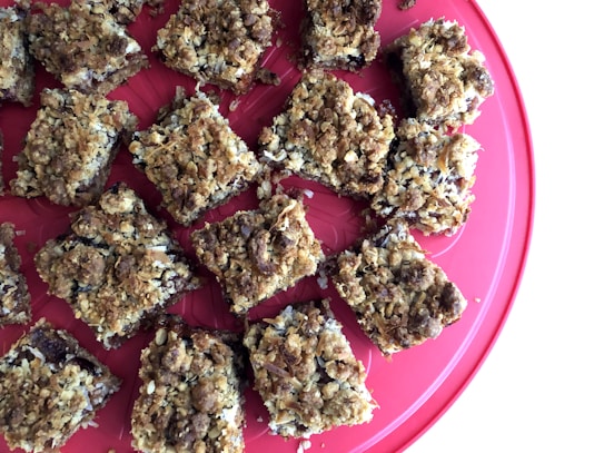 Several pieces of crumbly, oat-topped dessert bars arranged on a round, red plate. The texture appears crunchy with visible flakes and possible chocolate or fruit filling.