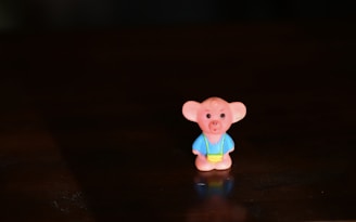 A small toy figure with a cartoonish appearance, resembling a bear or similar animal, wearing a blue outfit and a yellow accessory. It is placed on a dark surface, with soft lighting that highlights its features and casts a slight shadow.