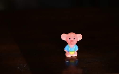 A small toy figure with a cartoonish appearance, resembling a bear or similar animal, wearing a blue outfit and a yellow accessory. It is placed on a dark surface, with soft lighting that highlights its features and casts a slight shadow.