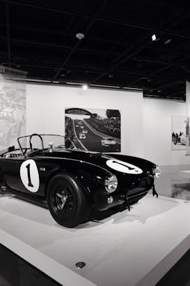 A classic black sports car with a number one displayed prominently on the hood and sides is showcased on a white platform in a gallery or museum setting. In the background, black and white photography depicting a racing event is displayed on the walls.
