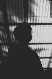 A silhouette of a person with short hair and glasses is captured against a backdrop of corrugated translucent material that allows light to filter through, casting linear patterns.