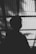 A silhouette of a person with short hair and glasses is captured against a backdrop of corrugated translucent material that allows light to filter through, casting linear patterns.