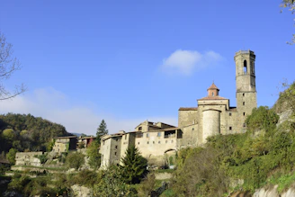 A picturesque medieval village is perched on a hill, characterized by its stone buildings and a prominent bell tower. The buildings are surrounded by lush greenery and trees, with a clear blue sky overhead.