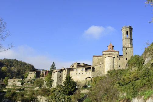 A picturesque medieval village is perched on a hill, characterized by its stone buildings and a prominent bell tower. The buildings are surrounded by lush greenery and trees, with a clear blue sky overhead.