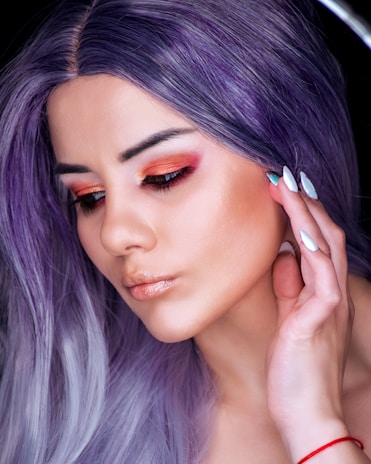 A person with long, purple hair and vibrant eye makeup featuring orange and red tones. The skin glows with a subtle shimmer, and there is attention to detail in the makeup application. Their nails are manicured with a combination of white and blue colors. A small red bracelet adorns the wrist.