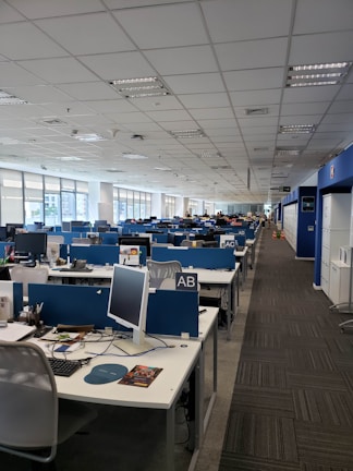 A spacious, modern office with rows of desks lined up in two aisles. The desks are equipped with computers, stationery, and other office supplies, organized neatly. Blue dividers separate the desks, and there are ergonomic office chairs. The ceiling is lined with fluorescent lights, and large windows on the left side allow natural light to enter. The overall atmosphere is quiet and orderly.