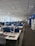 A spacious, modern office with rows of desks lined up in two aisles. The desks are equipped with computers, stationery, and other office supplies, organized neatly. Blue dividers separate the desks, and there are ergonomic office chairs. The ceiling is lined with fluorescent lights, and large windows on the left side allow natural light to enter. The overall atmosphere is quiet and orderly.