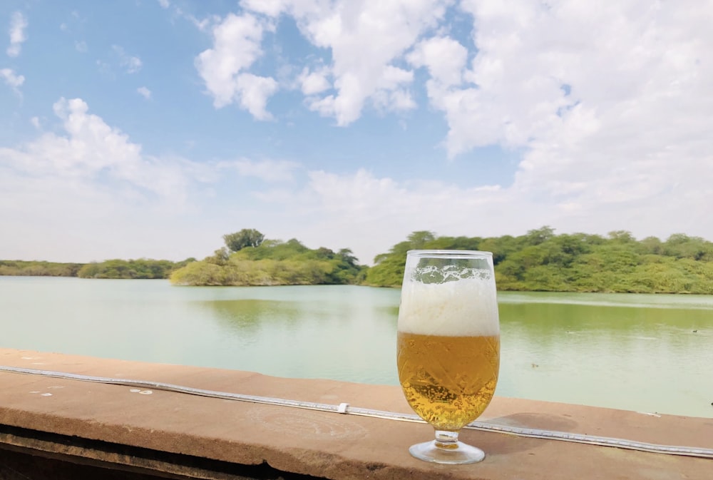 clear drinking glass with beer on brown wooden table near body of water during daytime