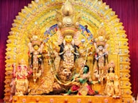 A richly decorated and colorful Durga Puja pandal displaying the goddess Durga with multiple arms, riding a lion, surrounded by other deities and figures adorned in vibrant garments and ornaments. The backdrop is intricately designed with golden and multicolored embellishments, creating a festive and majestic atmosphere.